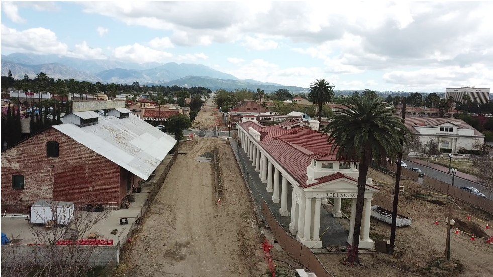 A dirt railway track runs between historic brick and white-columned buildings in Redlands, California, with mountains visible in the background.