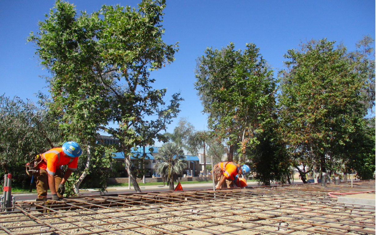 Two construction workers in orange shirts and blue helmets install steel rebar on a construction site surrounded by trees under a clear blue sky.