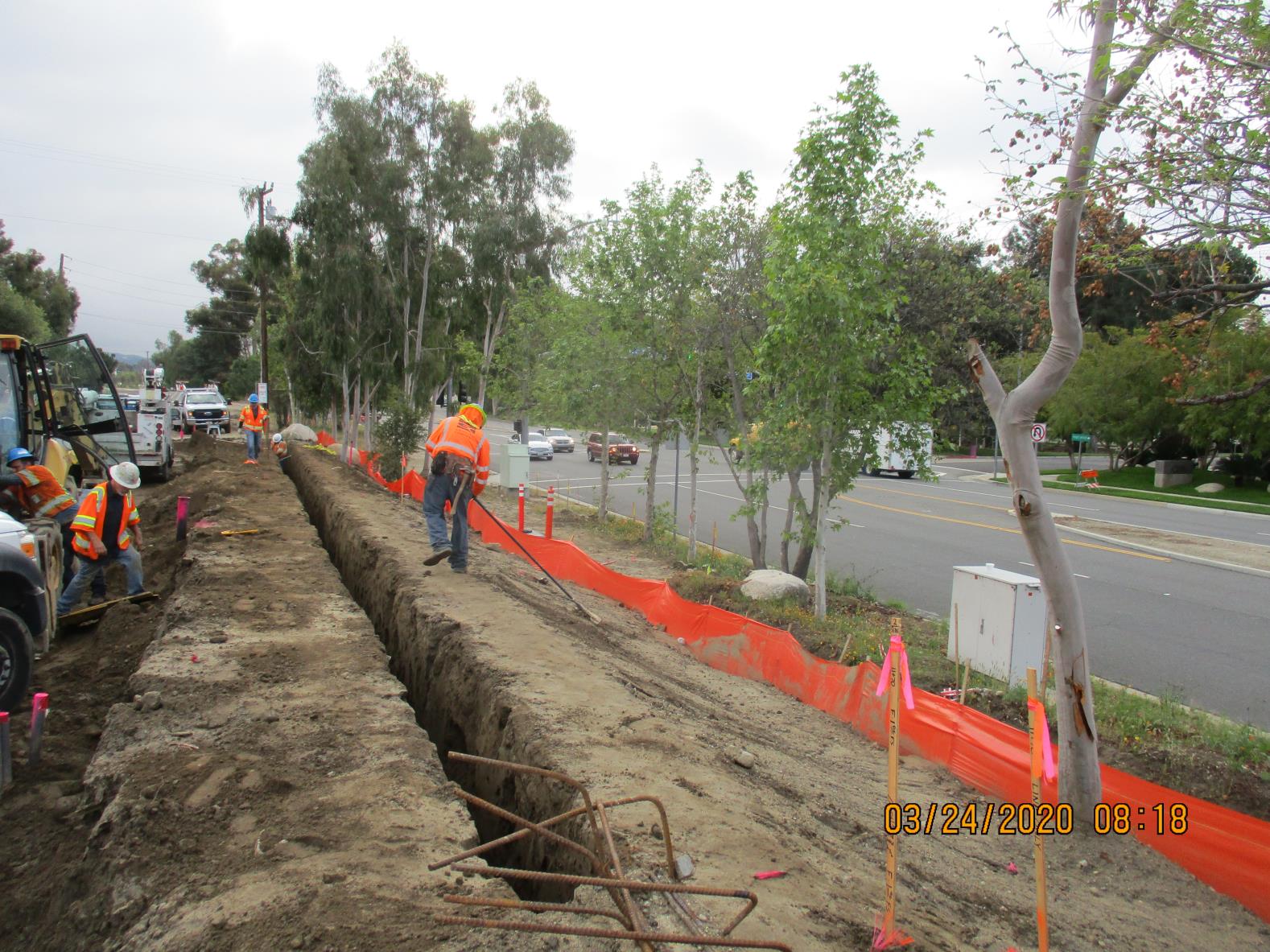 Construction workers in safety vests stand near a long trench beside a road lined with trees, with orange safety barriers and equipment visible.