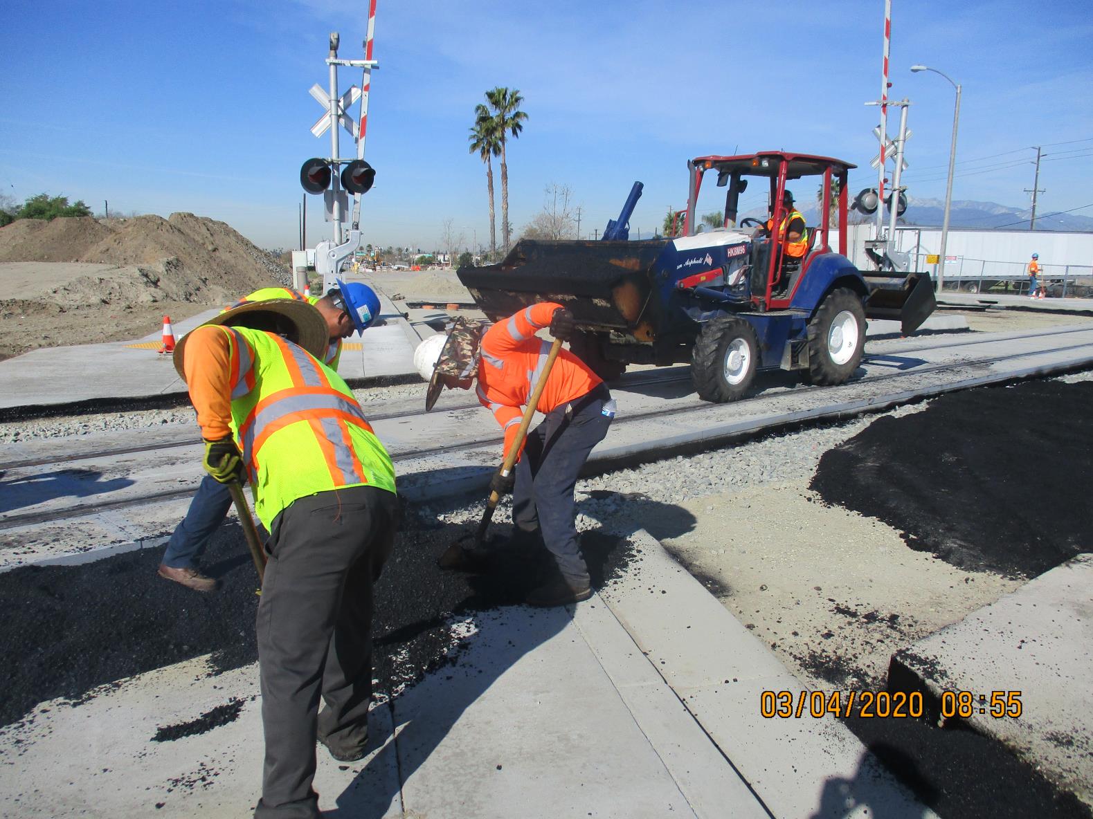 Construction workers in safety vests spread asphalt on a railway crossing as a tractor delivers more material; railroad signals and palm trees are visible in the background.