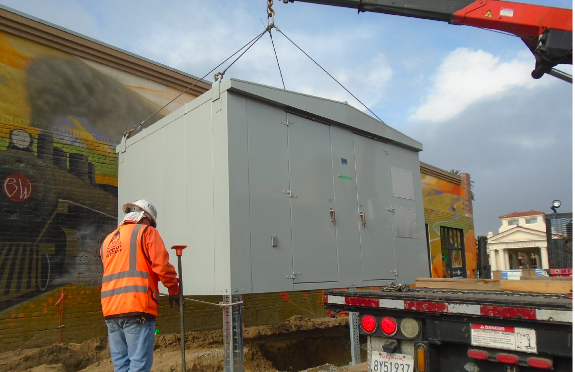 A worker in a safety vest guides a large metal utility box being lifted by a crane from a flatbed truck at a construction site near a mural.