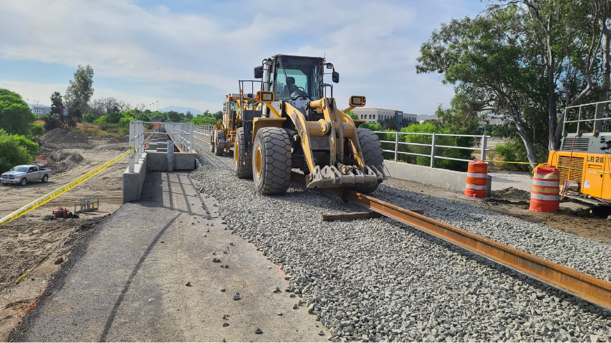 A front-end loader places a steel rail on a gravel railway bed during track construction; traffic barrels and safety barriers are visible nearby.