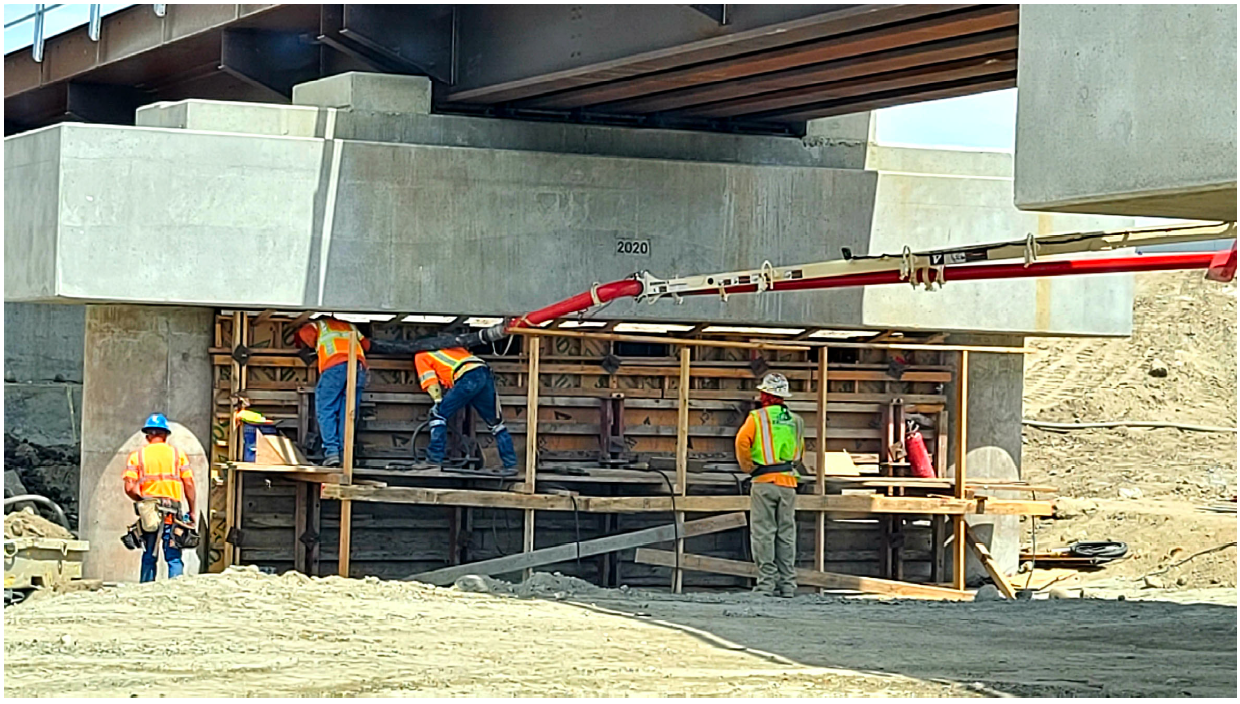 Five construction workers in safety gear work on scaffolding under a concrete bridge, with equipment and materials visible around them at the job site.