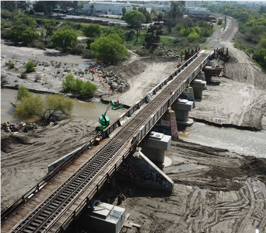 Aerial view of a railway bridge under construction over a river, with construction equipment and workers present on site.