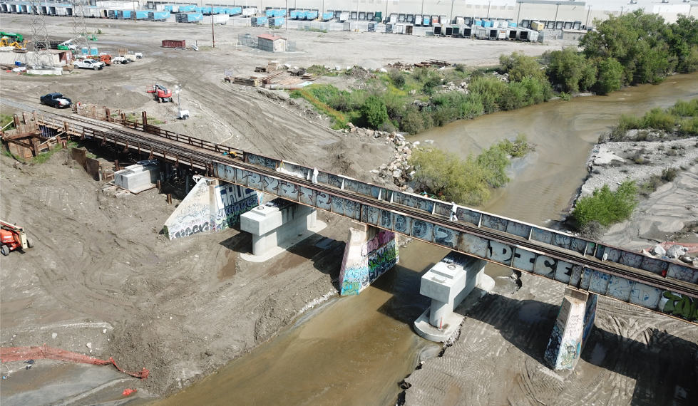 A rusted rail bridge with graffiti crosses a shallow river in an industrial area with construction equipment and buildings in the background.