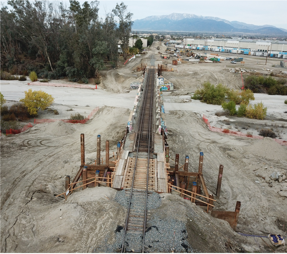 A railway bridge under construction spans a dry, sandy area with construction materials and equipment visible nearby; industrial buildings and mountains are in the background.