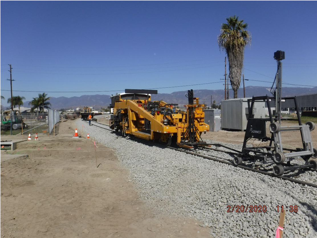 A yellow rail track construction machine works on laying new railroad tracks on a gravel bed at a construction site under a clear sky.