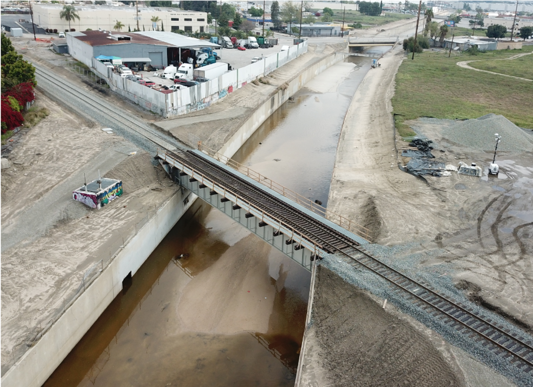 A single-track railway bridge crosses over a narrow concrete-lined canal with low water, surrounded by industrial buildings and open land.