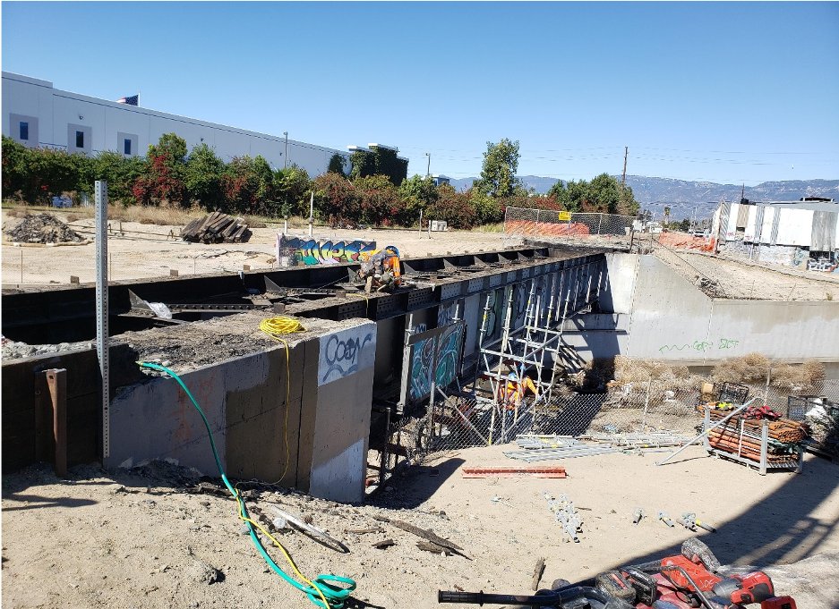 A construction site with a partially built concrete structure, scattered equipment, fencing, and graffiti on walls, under a clear blue sky.