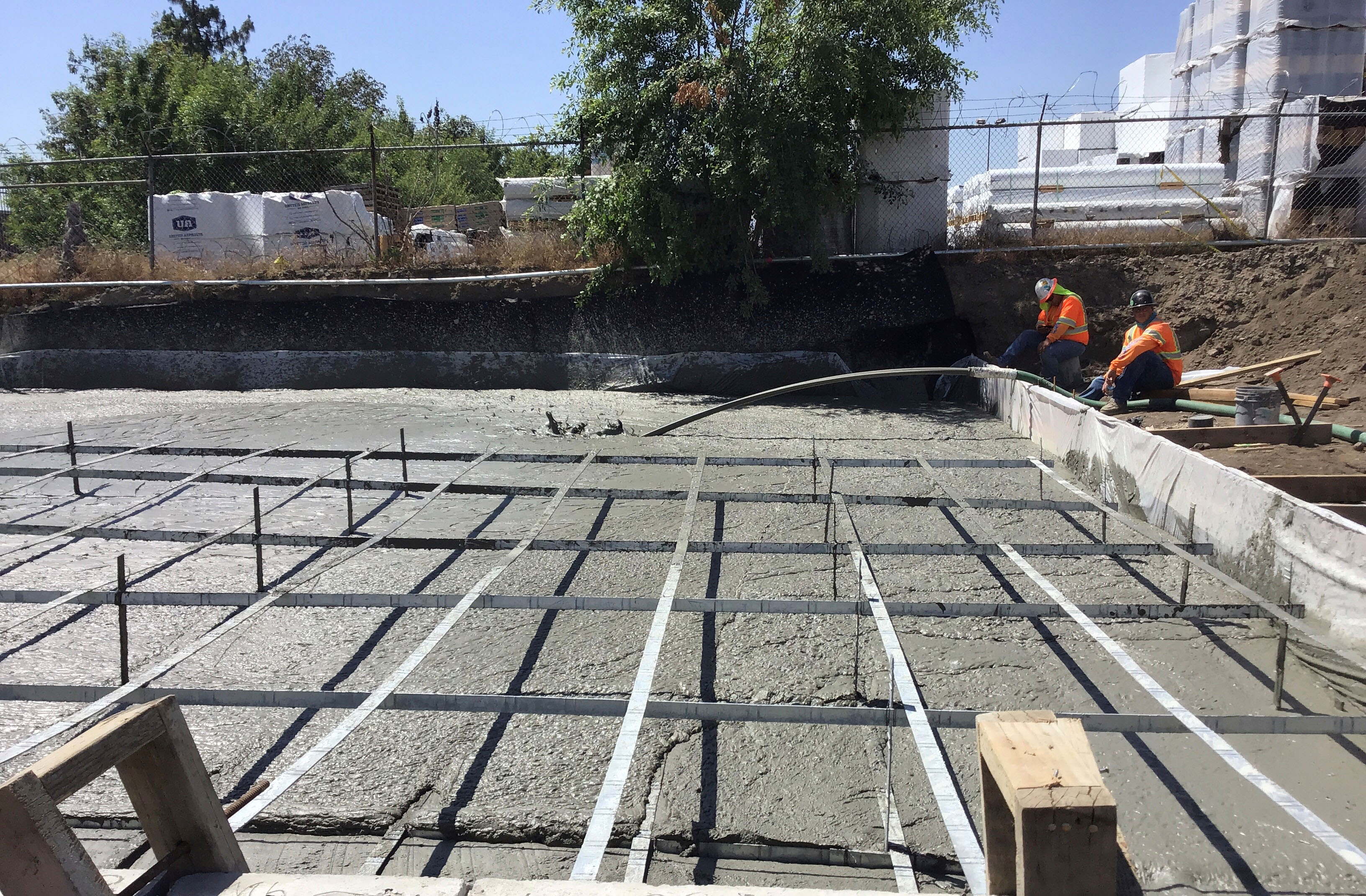 Two construction workers are smoothing wet concrete in a rebar-reinforced foundation at an outdoor construction site, with trees and materials visible in the background.