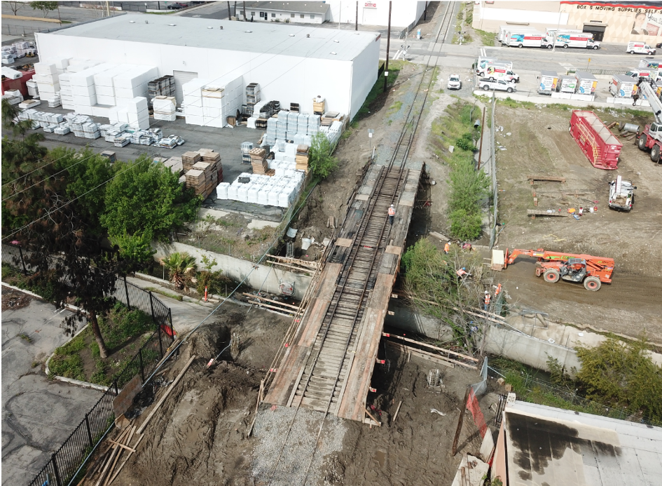 Aerial view of a construction site with a railway bridge over a road, surrounded by industrial buildings and construction equipment.