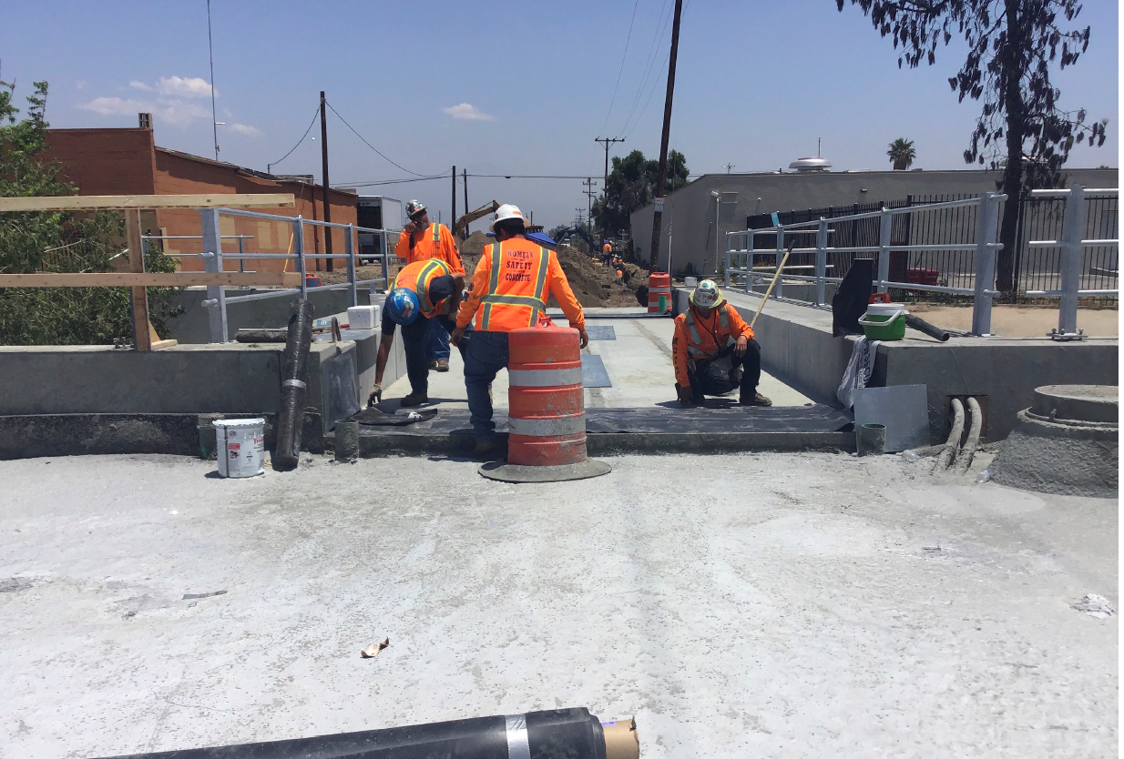 Four construction workers in safety gear work on a concrete bridge under clear skies, with equipment and barriers visible around the site.