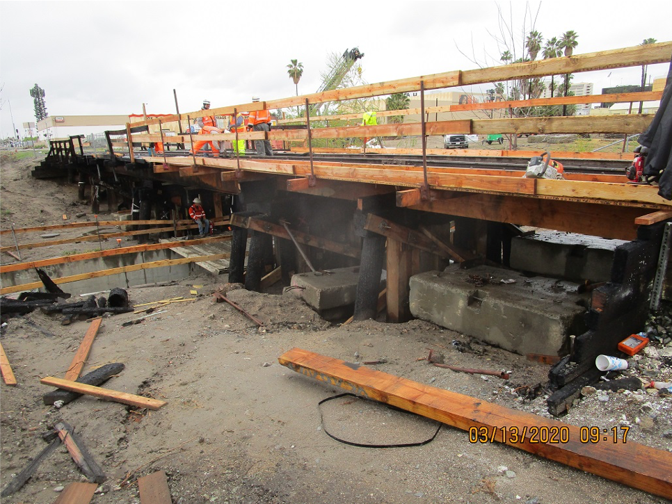 Wooden bridge under construction with workers and equipment present; exposed beams and support structures are visible beneath the bridge.