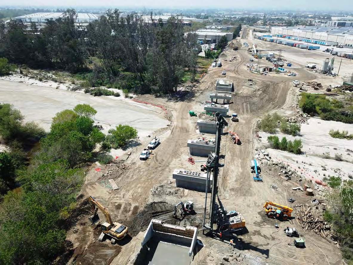 Aerial view of a construction site with heavy machinery, vehicles, and large concrete structures set along a cleared dirt area next to trees and industrial buildings.