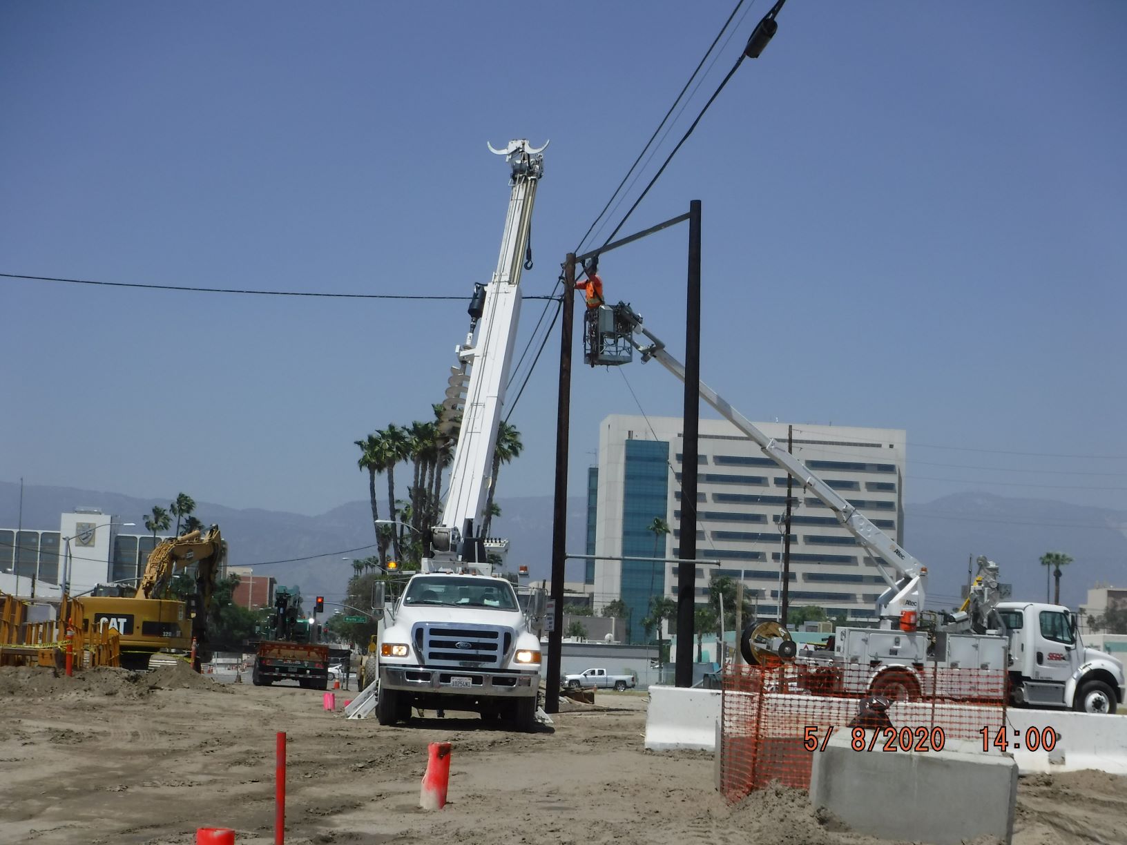 Construction site with cranes and workers installing utility poles; buildings and construction equipment are visible in the background; photo dated 05/07/2020 at 14:00.