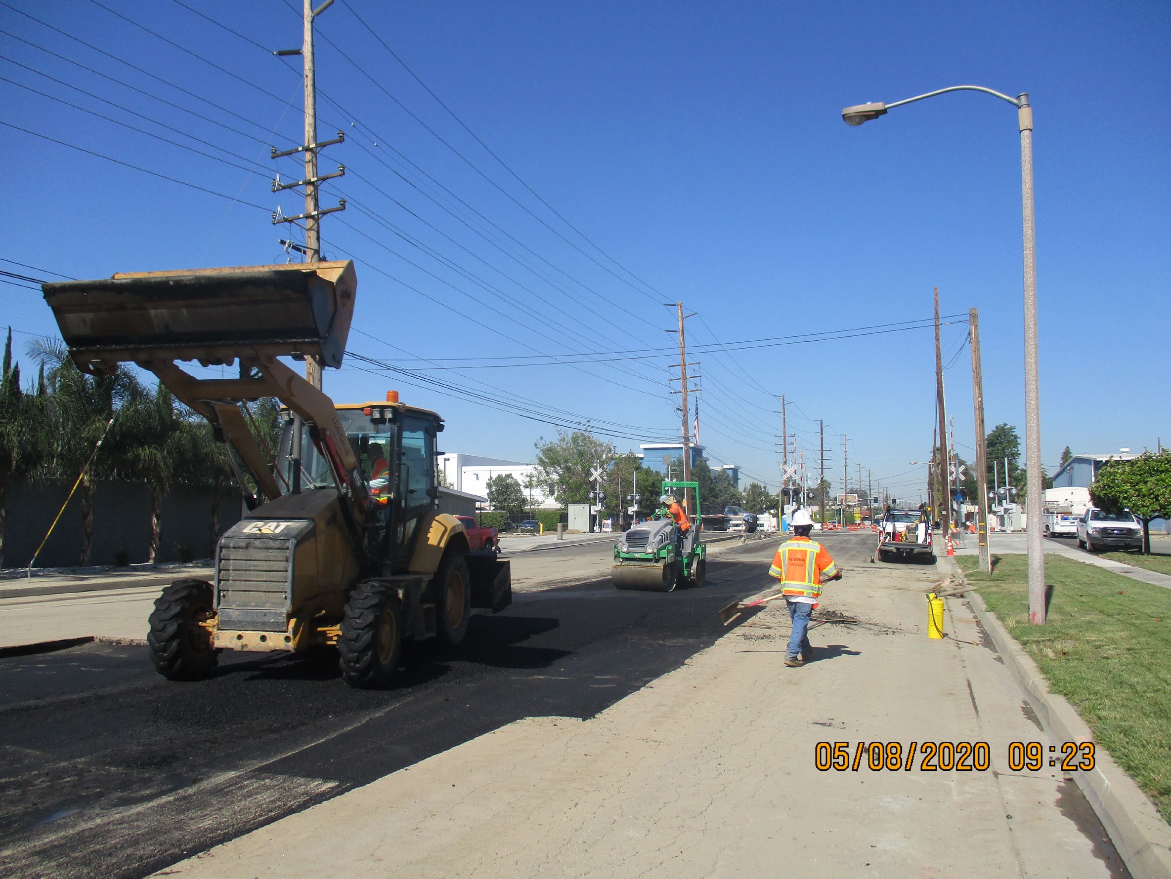 Construction workers repave a multi-lane road using heavy machinery under clear skies. One worker stands near a backhoe loader. The date stamp reads 05/08/2020 09:23.