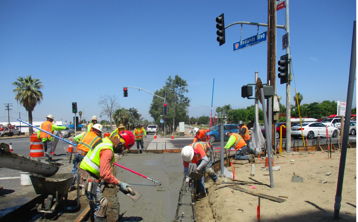 Construction workers in safety vests pour and smooth concrete at a roadside under clear blue skies near a traffic intersection with cars and traffic signals visible.