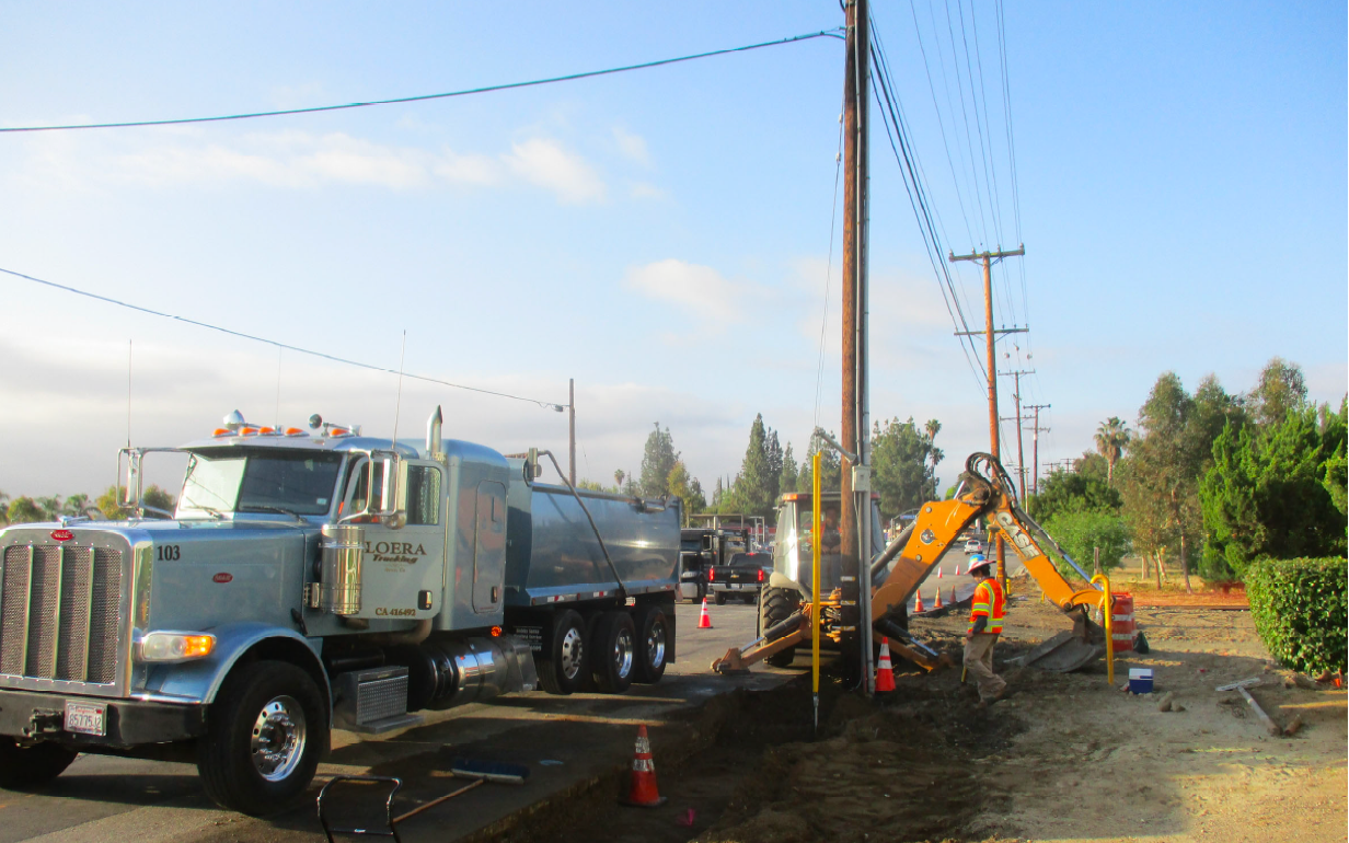 A construction crew uses an excavator to dig near power lines on a roadside while a dump truck is parked nearby; traffic cones are set up for safety.