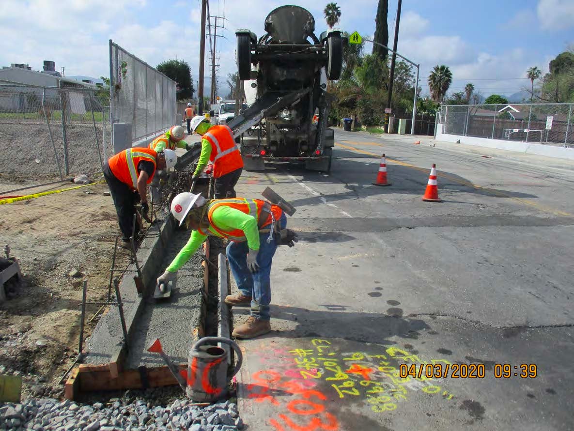 Construction workers pour and level concrete on a road as a cement truck dispenses material; traffic cones block off the work area.
