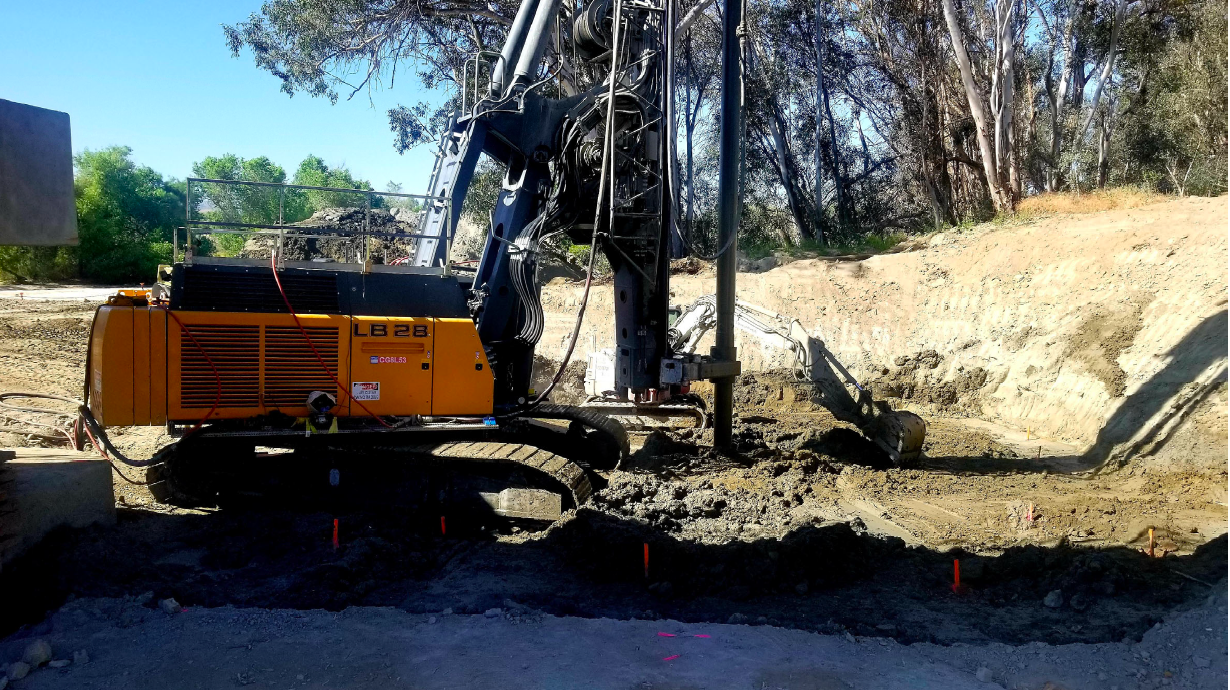 A large yellow drilling machine is positioned on a construction site with dirt, marked stakes, and trees in the background.