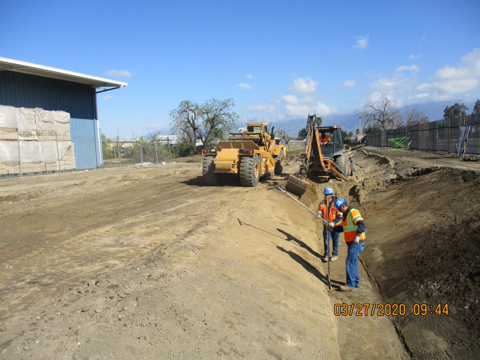 Two construction workers in safety gear stand near an excavated trench, while heavy machinery operates in the background on a construction site.