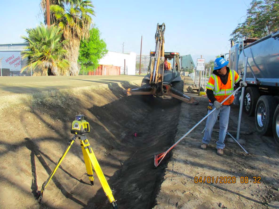 A construction worker smooths a dirt trench with a rake near an excavator as a laser level stands nearby. The scene is outdoors with construction vehicles and palm trees in the background.