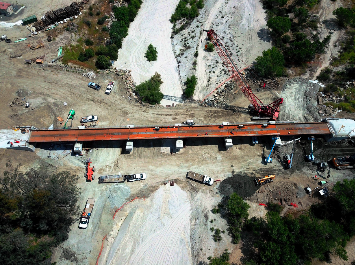 Aerial view of a construction site with a bridge spanning a dry, sandy area. Trucks, construction vehicles, and equipment are positioned around the bridge and site.