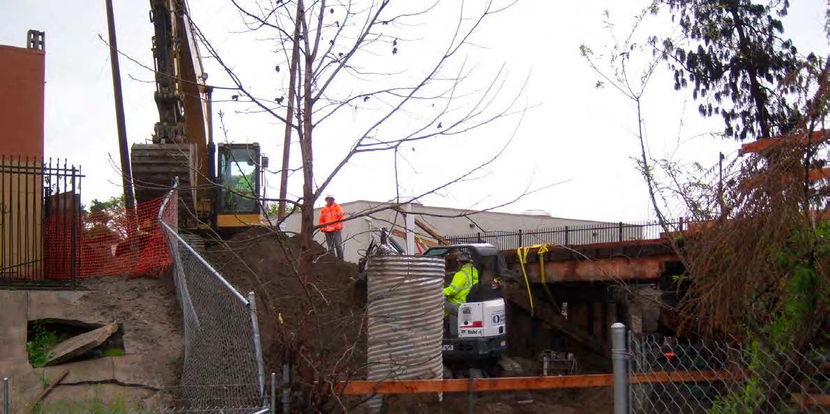 Construction workers operate heavy machinery near a fenced area, working on an embankment and bridge structure with trees and buildings nearby.