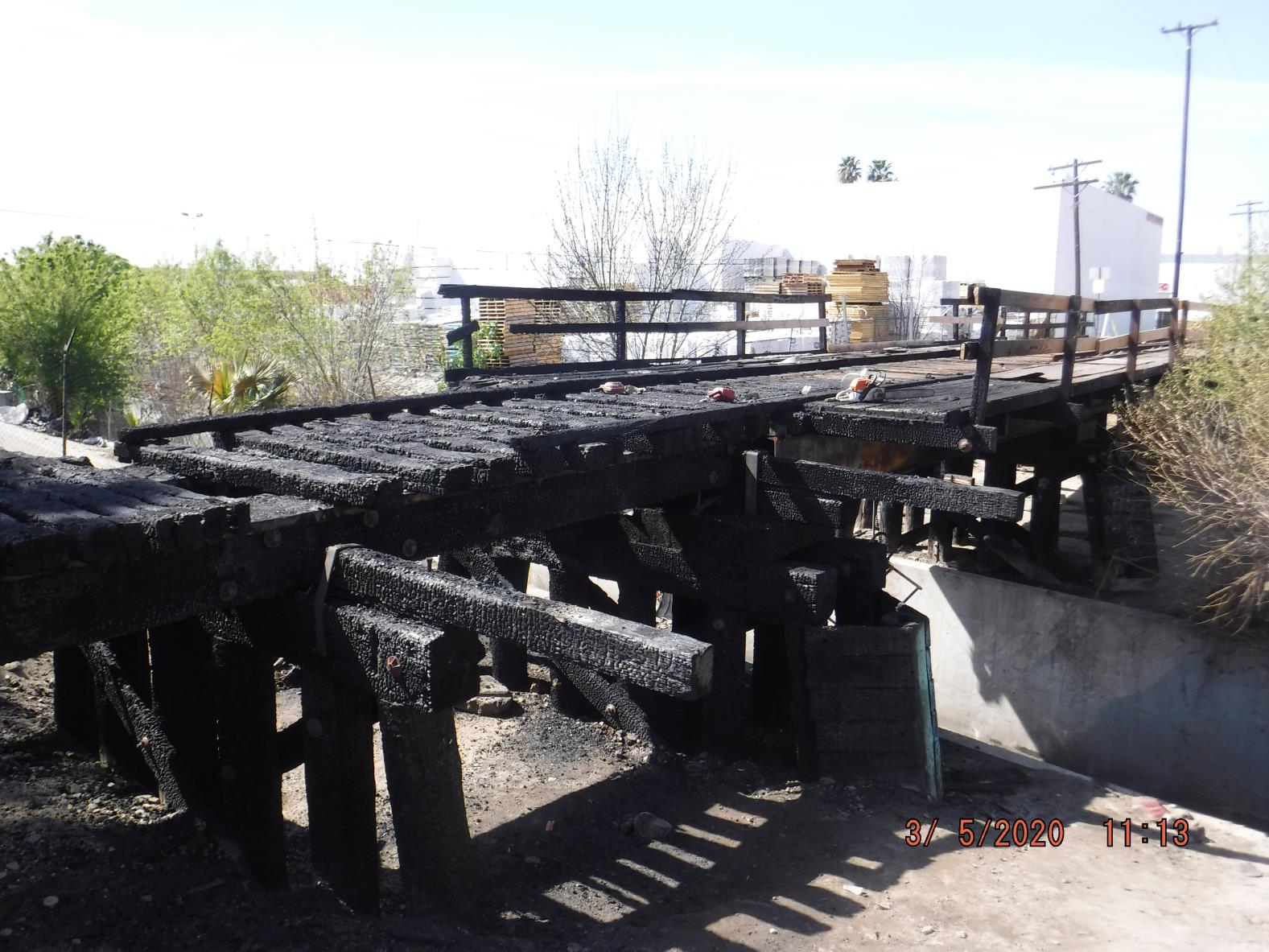 A charred, damaged wooden bridge with blackened beams and missing planks, surrounded by sparse vegetation and urban structures in the background.