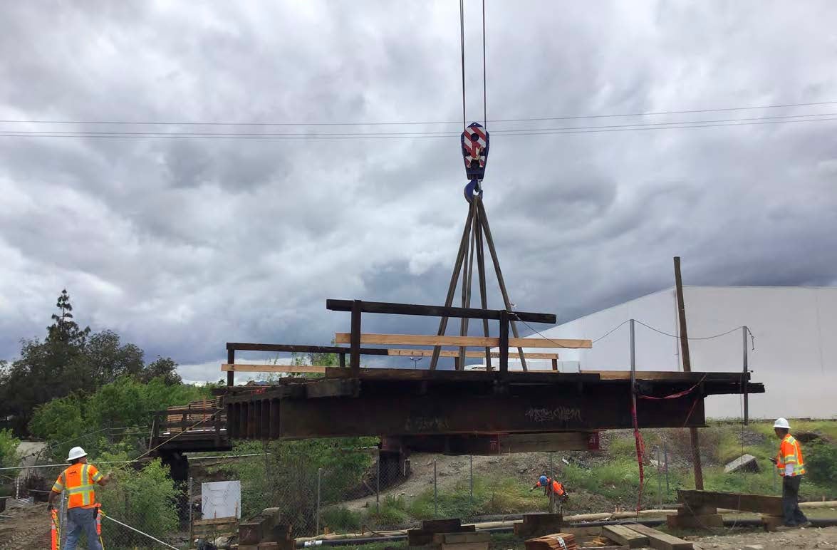 A crane lifts a large steel structure at a construction site while workers in safety vests and helmets monitor the operation under a cloudy sky.