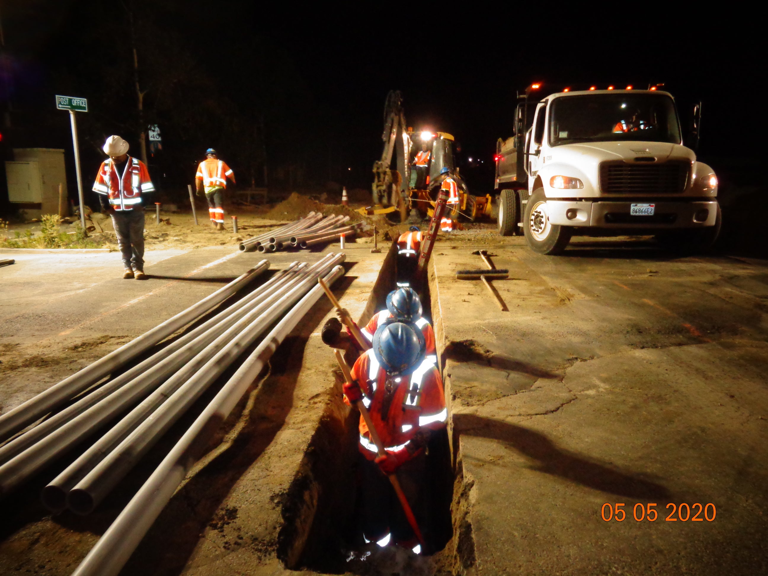 Construction crew works at night installing pipes in a trench along a road, with safety gear, machinery, and trucks visible. Photo dated May 5, 2020.