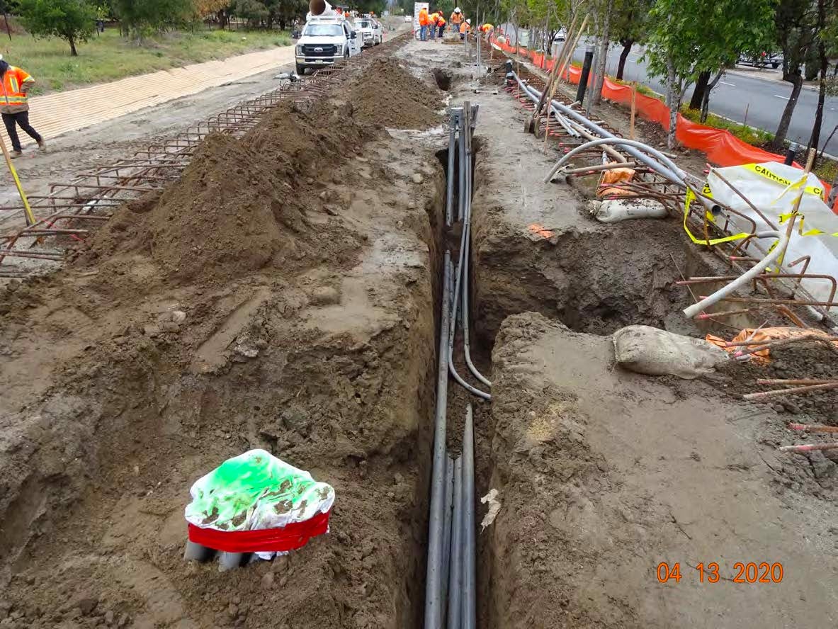 Construction workers are installing underground utility cables in a long, open trench beside a road lined with trees and orange safety barriers.