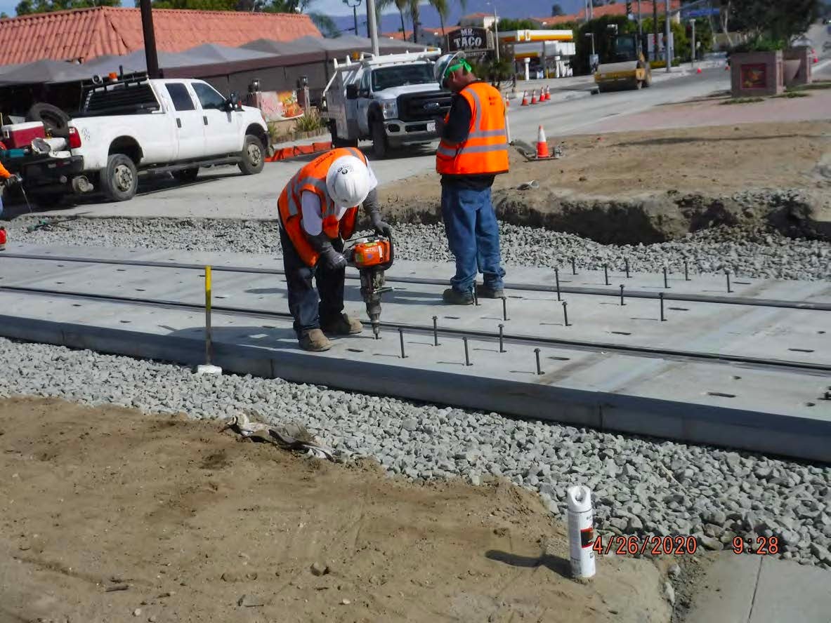 Workers in safety gear use power tools to install bolts on a concrete track section at a construction site with trucks and equipment in the background.