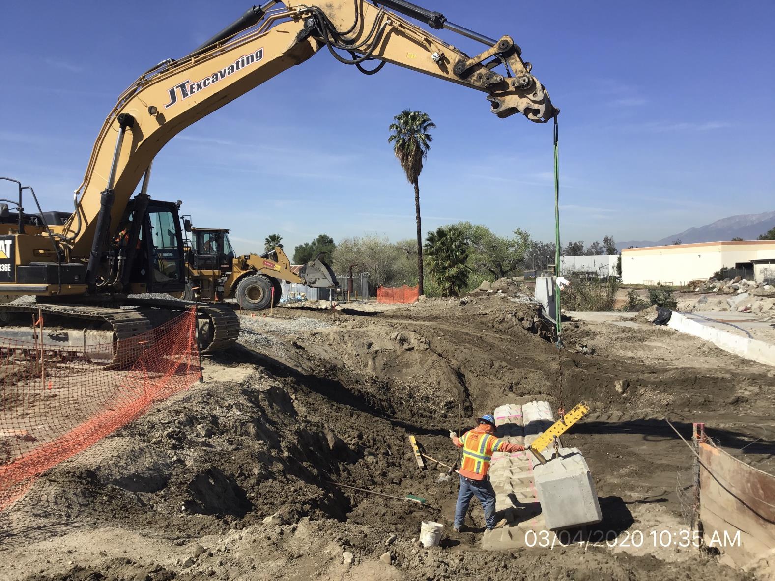 A construction worker guides a large concrete block being lifted by an excavator at an outdoor construction site with dirt, fencing, and nearby palm trees.
