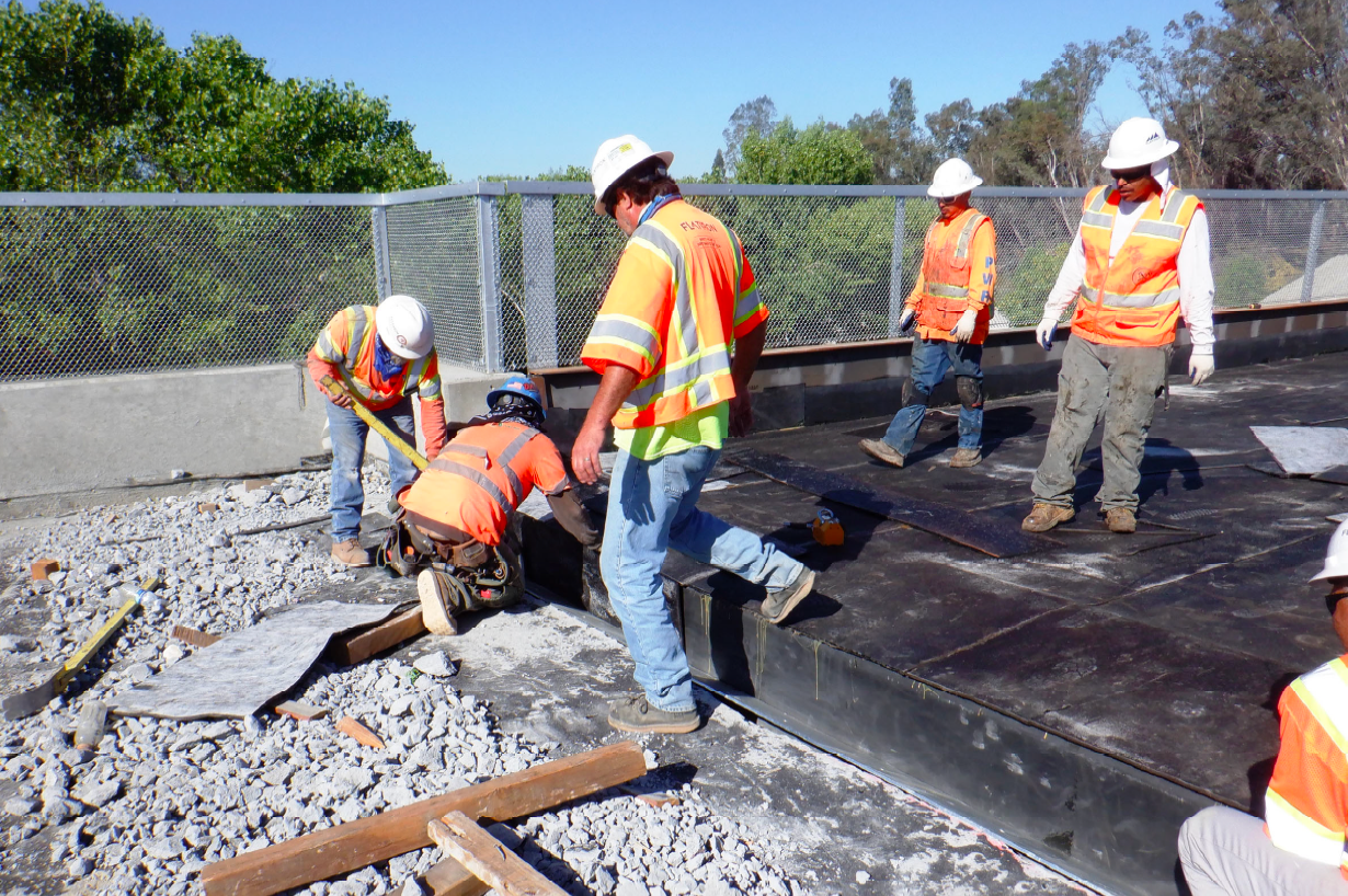 Construction workers in safety vests and helmets install materials on a paved surface outdoors, surrounded by gravel and fencing under a clear sky.