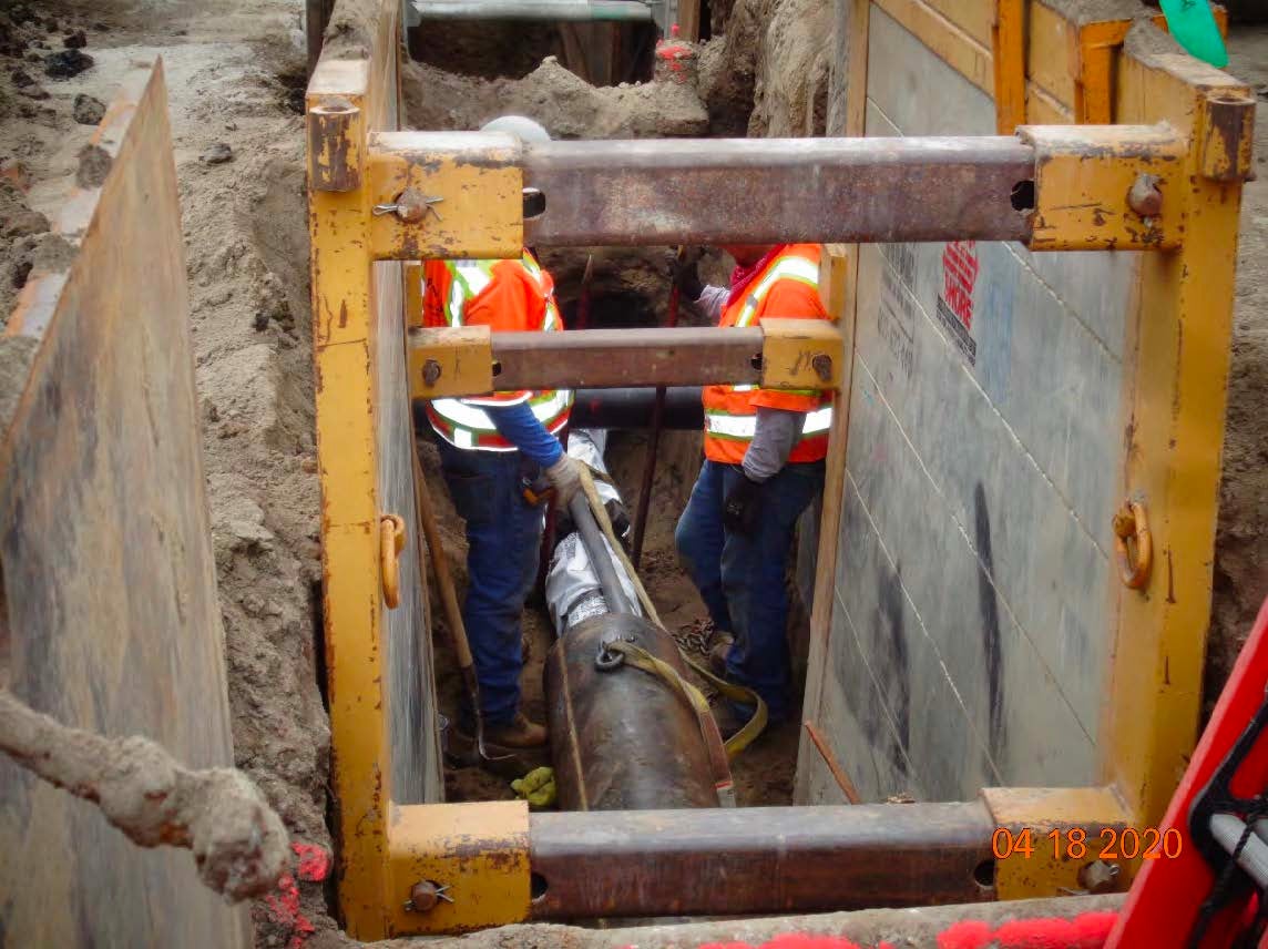 Two workers in safety vests install or inspect a large pipe in a reinforced trench at a construction site.