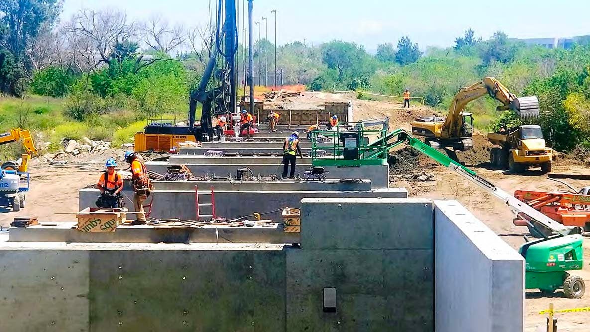 Construction workers operate machinery and work on a bridge or road structure at an outdoor construction site surrounded by greenery.