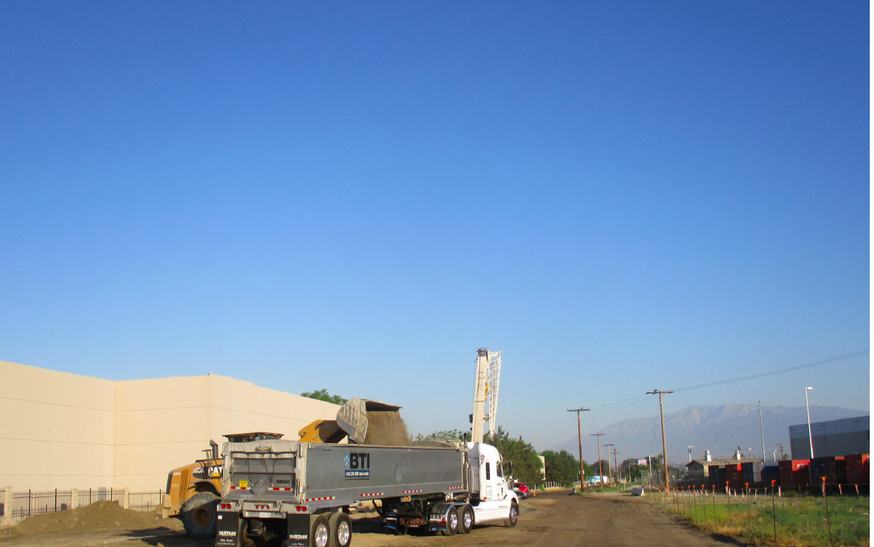 A construction site with a dump truck unloading dirt into a BTI company truck on a dirt road, with a crane and mountains in the background under a clear blue sky.