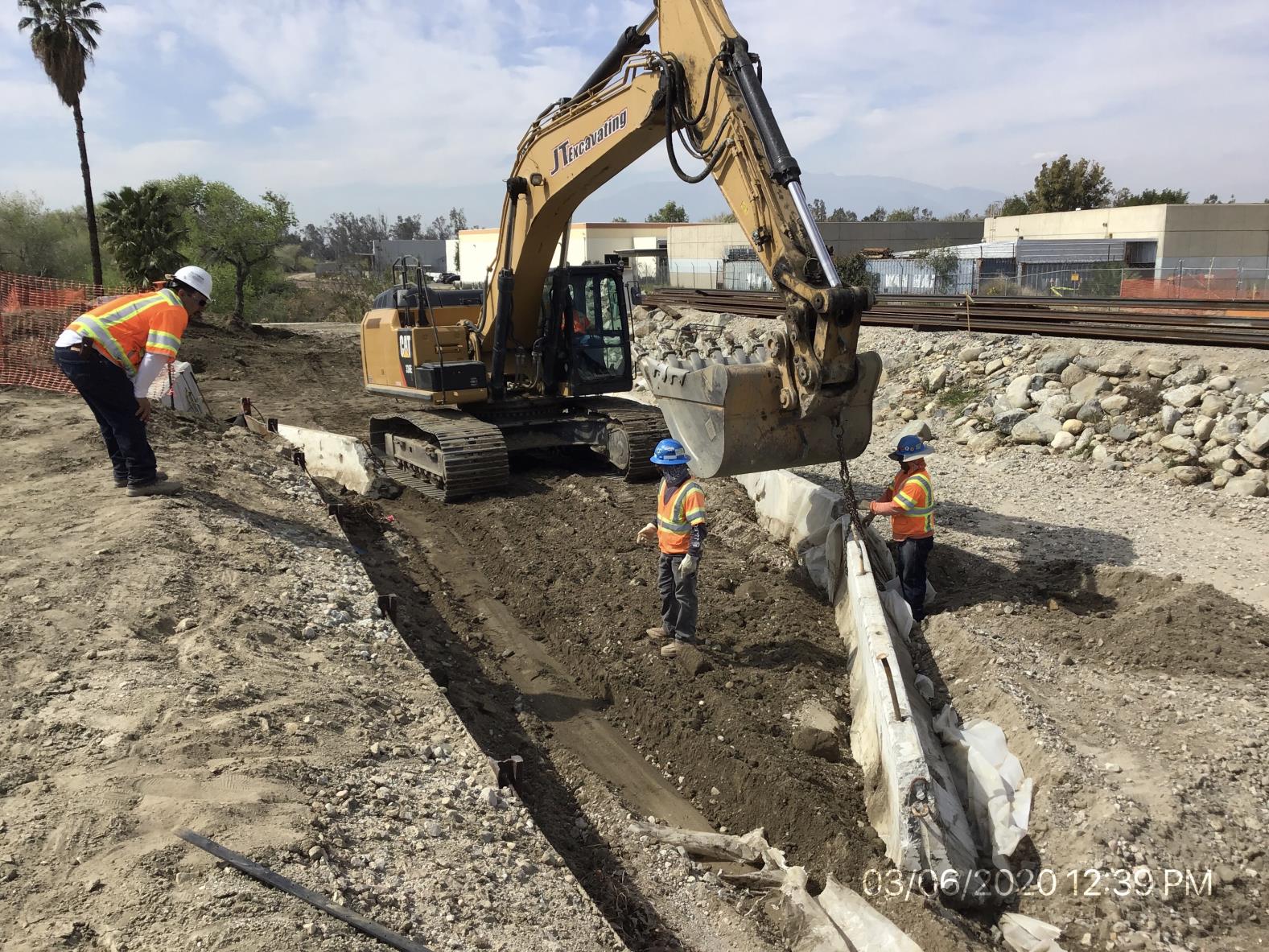 Three construction workers in safety gear operate and guide an excavator digging near a rail track; soil is being moved beside a temporary retaining barrier.