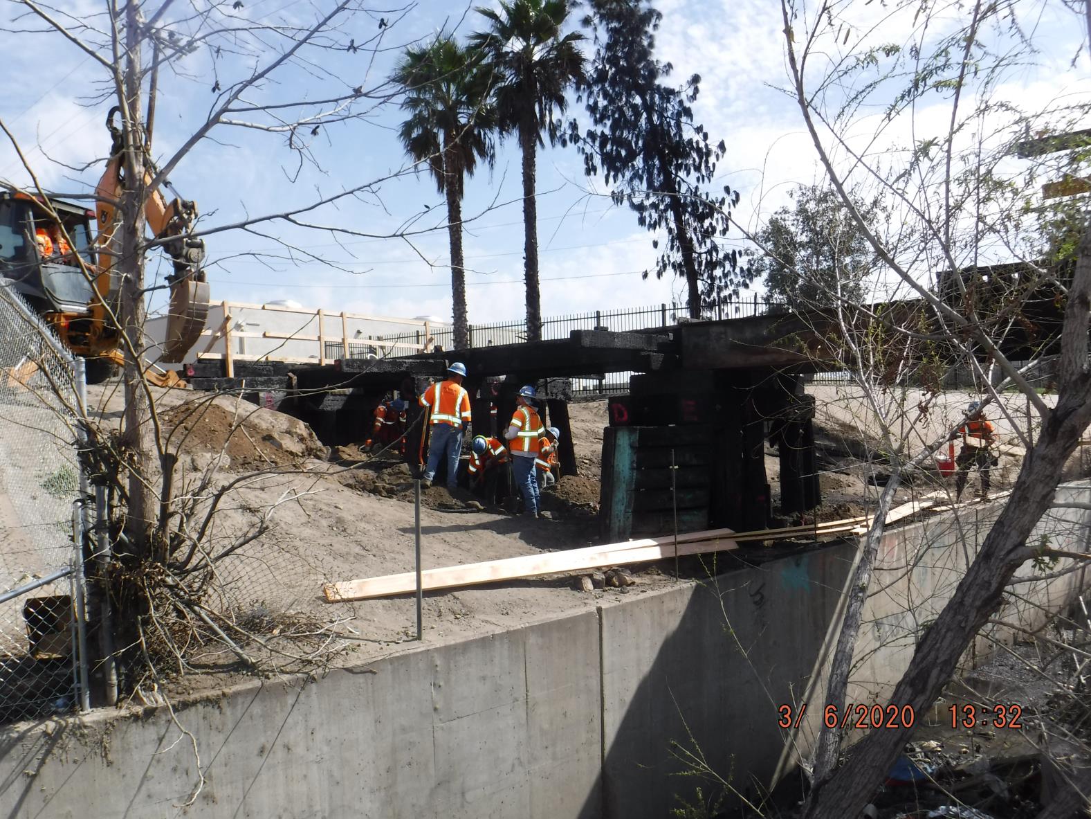 Construction workers in safety vests work beneath a partially collapsed bridge near a dirt embankment, with construction equipment and palm trees in the background.