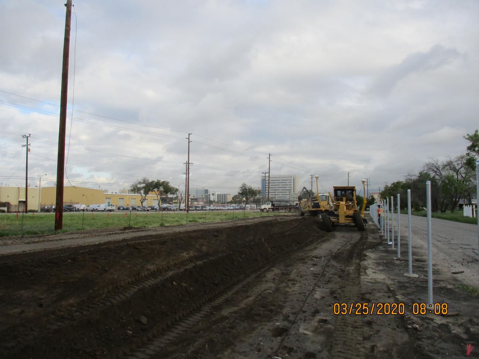 Construction site with bulldozers leveling dirt near a road; white poles installed along the roadside; overcast sky and city buildings in the background.