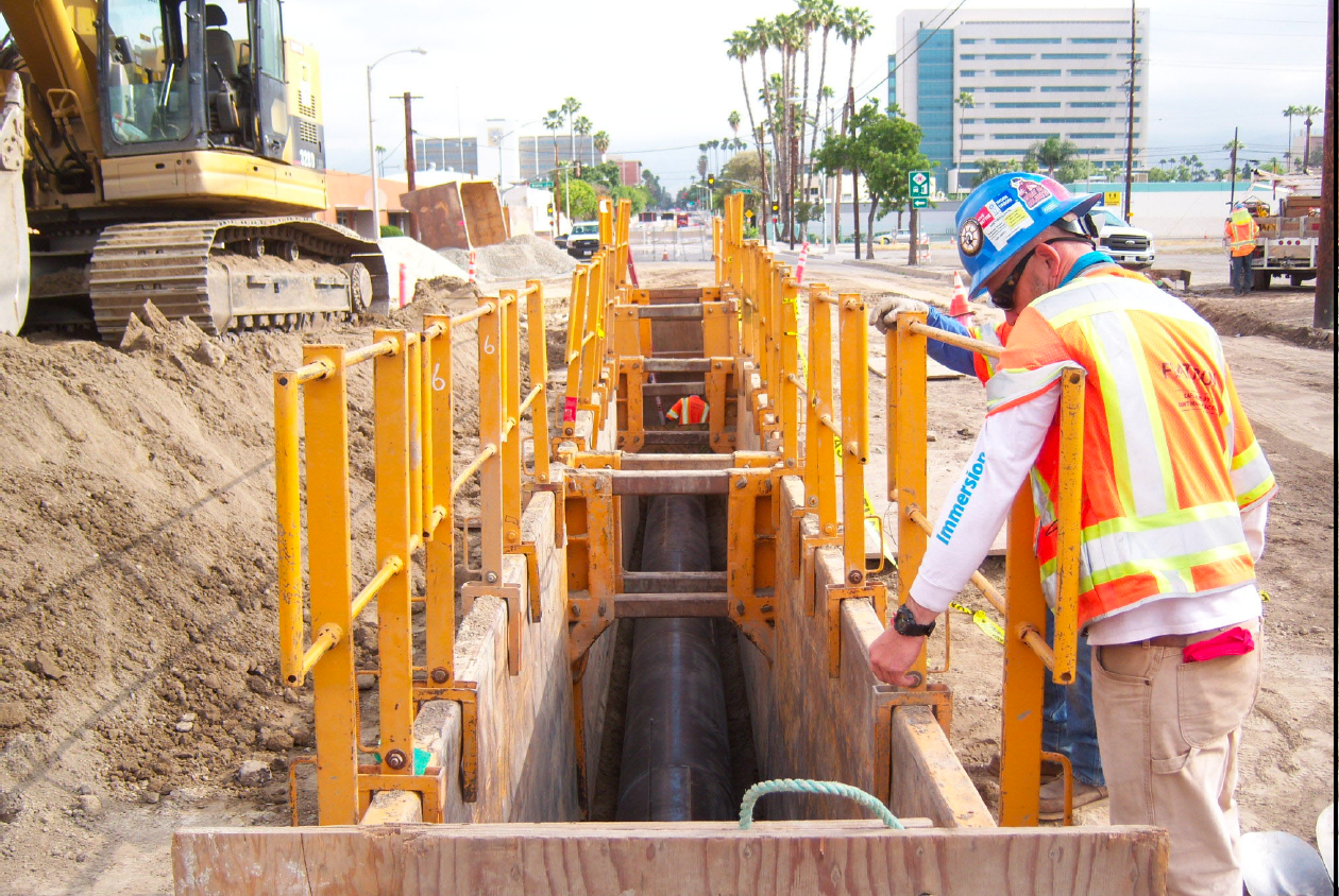 A construction worker inspects a trench reinforced with yellow barriers at a street worksite, with heavy machinery and buildings visible in the background.