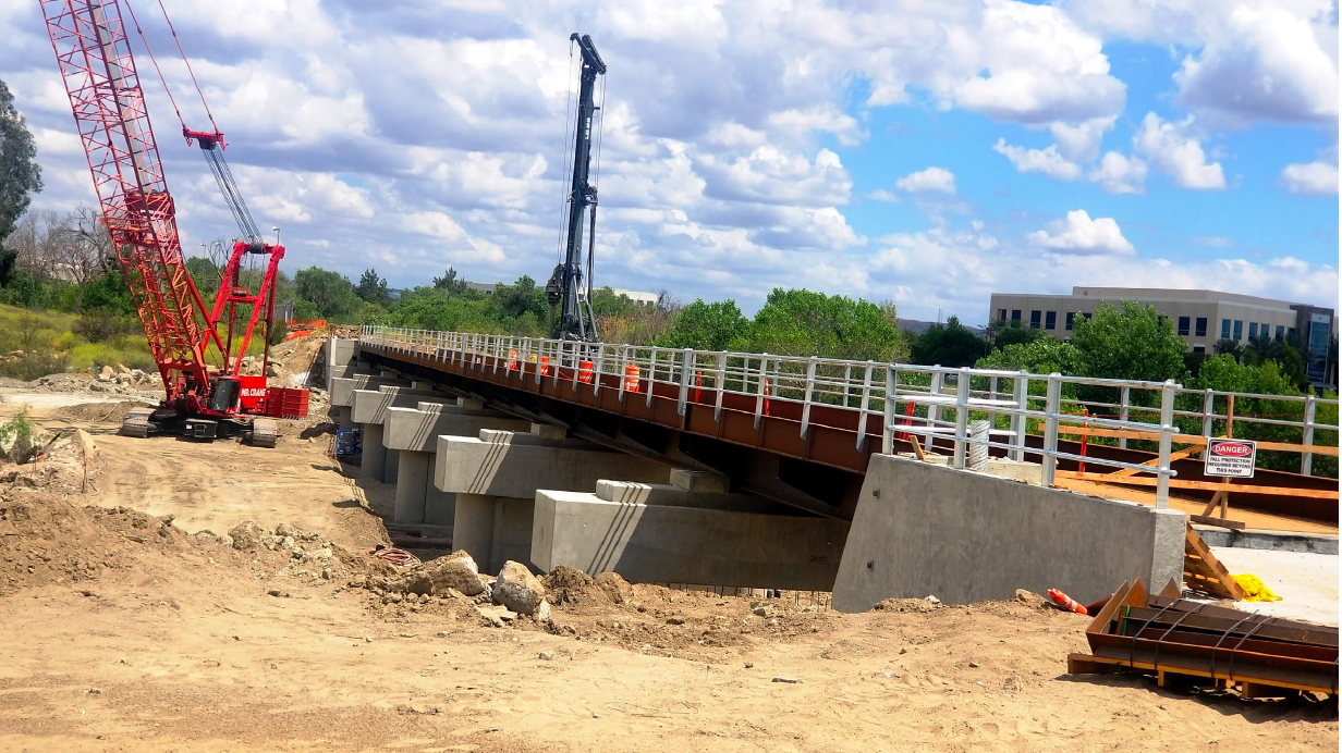 A partially constructed bridge with cranes and construction equipment on a dirt site under a partly cloudy sky.