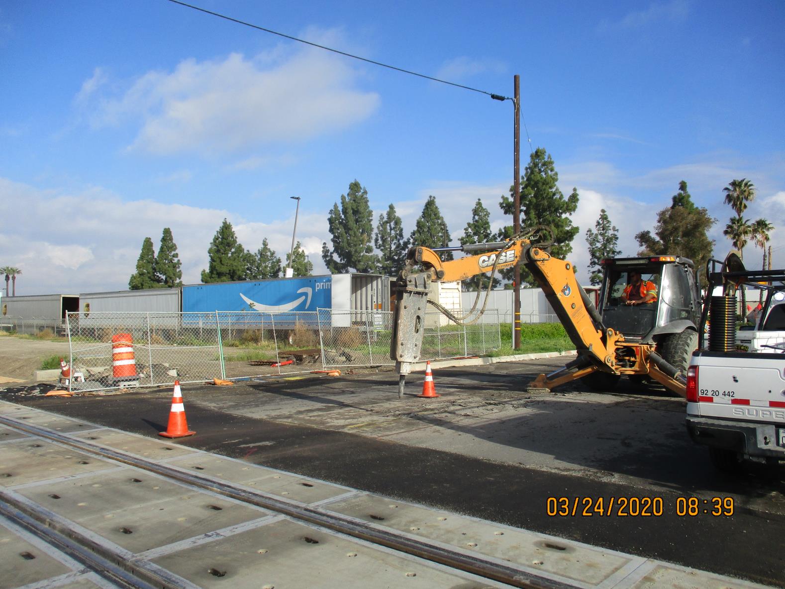 A construction vehicle breaks pavement near railroad tracks, with traffic cones and fencing marking the work area. Trees and an Amazon building are visible in the background.