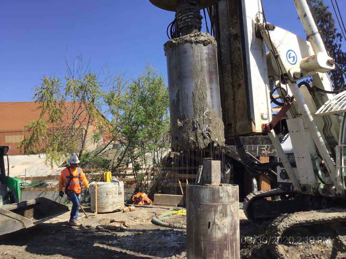 A construction worker stands near a drilling machine boring a large concrete shaft at an outdoor construction site.
