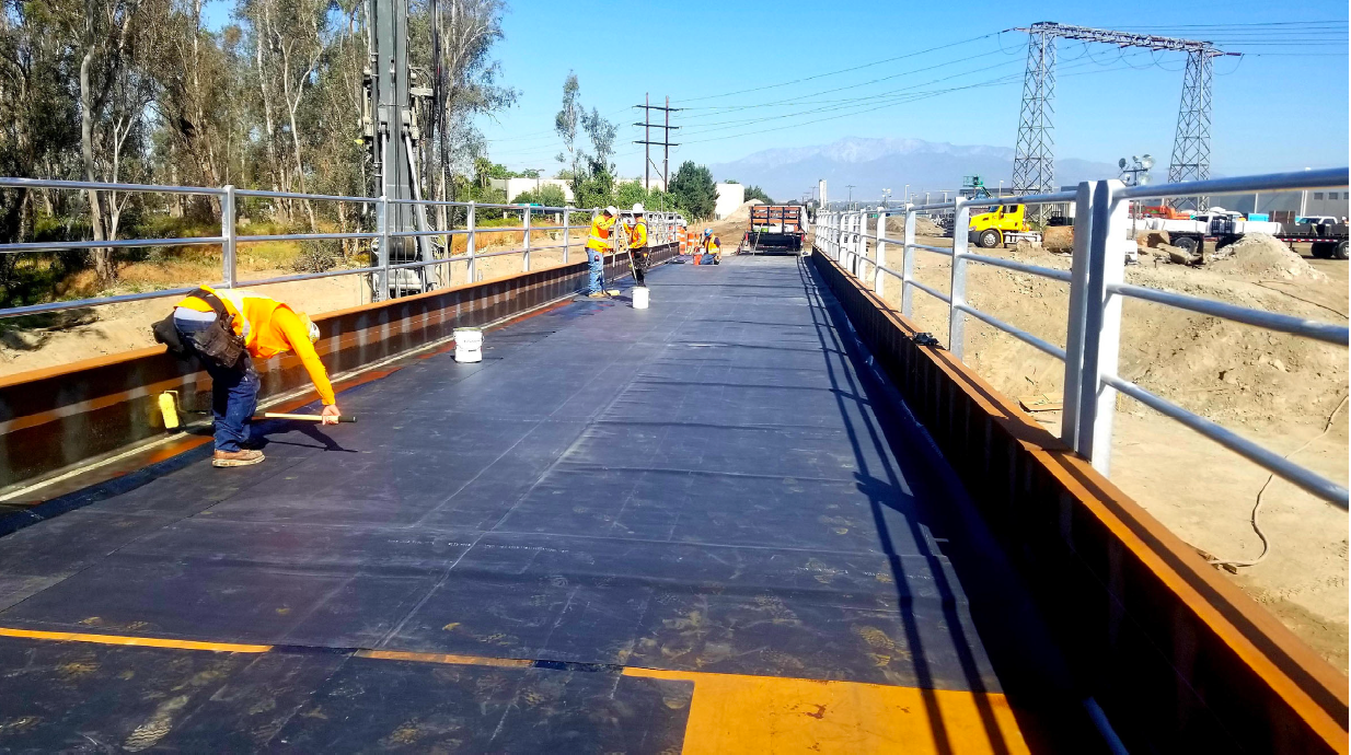 Workers in safety gear apply material to the surface of a bridge under construction, with construction vehicles and electrical towers visible in the background.