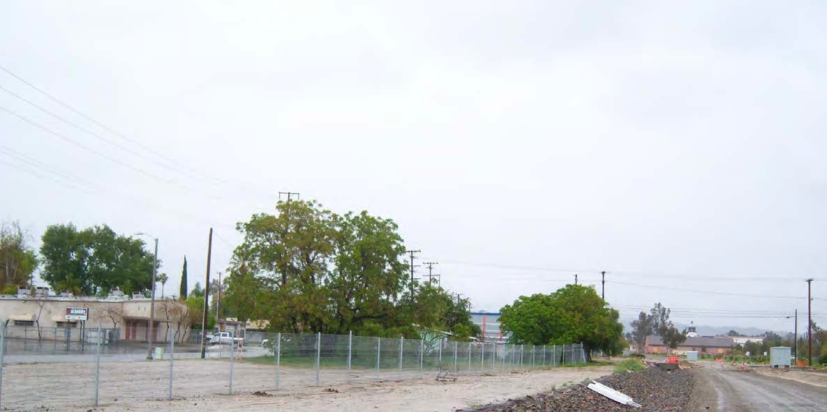 A chain-link fence separates a dirt lot from a row of trees and buildings under an overcast sky. Power lines and utility poles are visible in the background.