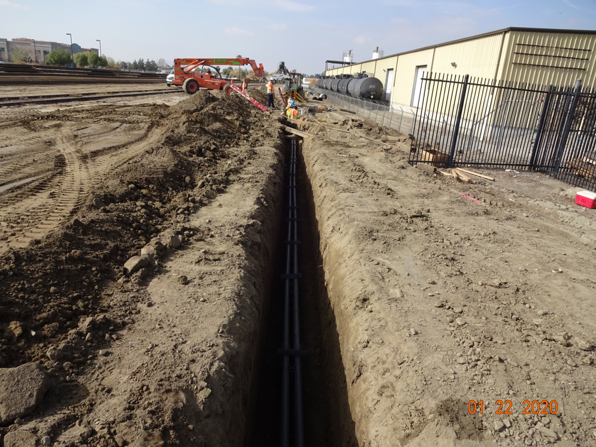Workers installing pipes in a long trench next to a building and fencing on a construction site, with equipment and materials visible in the background.