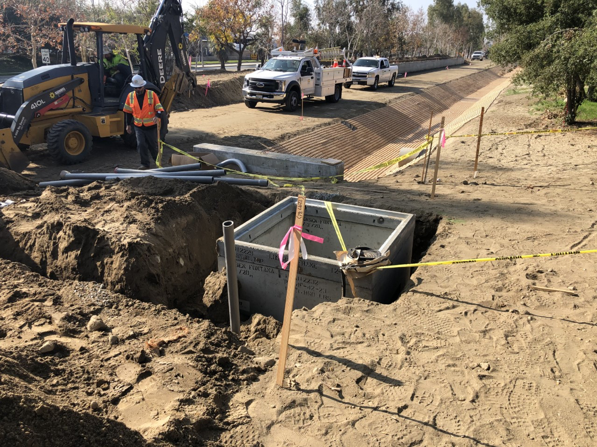 Construction workers and equipment at a roadside site with an open concrete utility box, caution tape, and exposed soil.