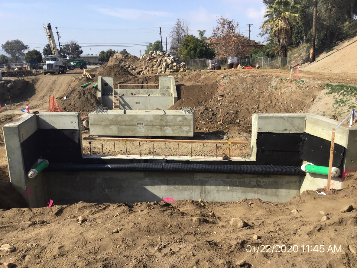 A construction site showing partially built concrete bridge abutments and retaining walls, with exposed pipes and soil around the structure.